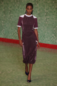 Female model walking a runway in a burgundy sequin midi dress with white shoulder panels and cuffs, against a green tiled wall background.