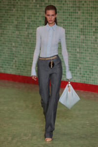 Model walks a runway in a pale blue sheer button-down blouse and dark gray trousers with a beige belt, carrying a pale blue bag against a green tiled backdrop.