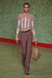Model walking a runway in a beige color-blocked blouse with white panels and brown trousers, carrying a beige handbag and wearing a brown belt in front of a green tiled backdrop.