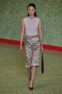 Model walking runway in a sleeveless white textured crop top and a high-waisted botanical print midi skirt, carrying a brown clutch.