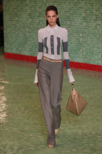 Model on a runway wearing a white striped button-down top with dark vertical panels, gray high-waisted trousers, a beige belt, and a tan handbag against a green tiled wall.