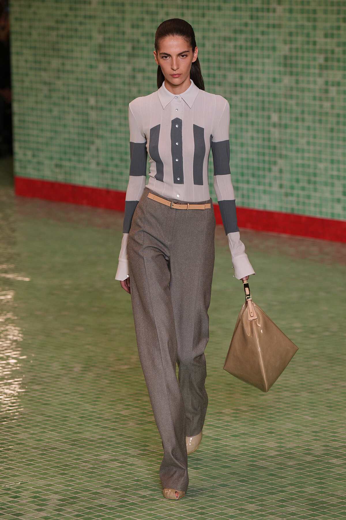 Model on a runway wearing a white striped button-down top with dark vertical panels, gray high-waisted trousers, a beige belt, and a tan handbag against a green tiled wall.
