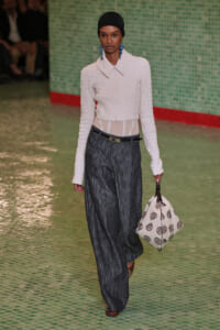 Model walking the runway in a white textured cropped jacket, wide dark trousers, and a black headscarf, accessorized with blue earrings and carrying a white patterned bag.
