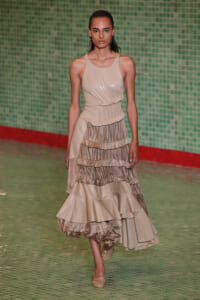 Model walks the runway in a beige sleeveless, tiered gown with a ruched waist and ruffled layers.