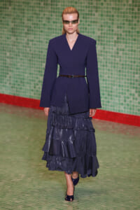 Runway model in a navy wrap blazer with a belted waist and a layered, ruffled navy skirt, wearing dark sunglasses.
