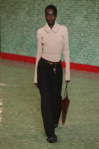 Model walks a runway in a cream textured knit top with a large collar and black wide‑leg pants, accessorized with a brown belt and a brown handbag.