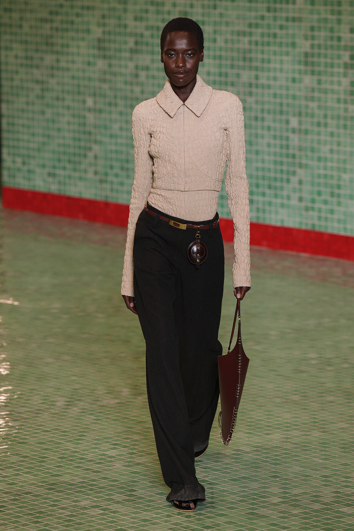Model walks a runway in a cream textured knit top with a large collar and black wide‑leg pants, accessorized with a brown belt and a brown handbag.