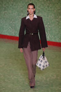 Model on a runway wearing a dark pinstripe blazer with a white collar, belt, pink wide-leg trousers, and a white bag with blue circular prints.