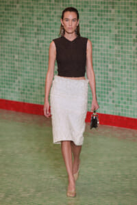 Model walking a runway in a black sleeveless textured crop top and white textured midi skirt, carrying a small metallic handbag.