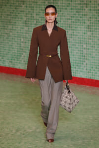 Model walks runway in a brown wrap blazer with a slim belt, paired with gray wide-leg trousers and brown open-toe heels, carrying a patterned tote and wearing orange sunglasses.