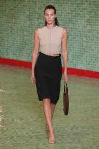 Model walking a runway in a beige textured sleeveless top and black knee-length skirt, carrying a brown handbag.