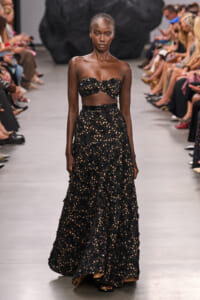 Model on a runway wearing a black floral lace two-piece outfit: strapless crop top and a high-waisted, full-length skirt with a belt.