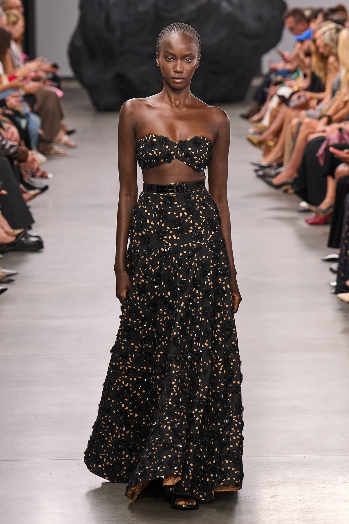 Model on a runway wearing a black floral lace two-piece outfit: strapless crop top and a high-waisted, full-length skirt with a belt.