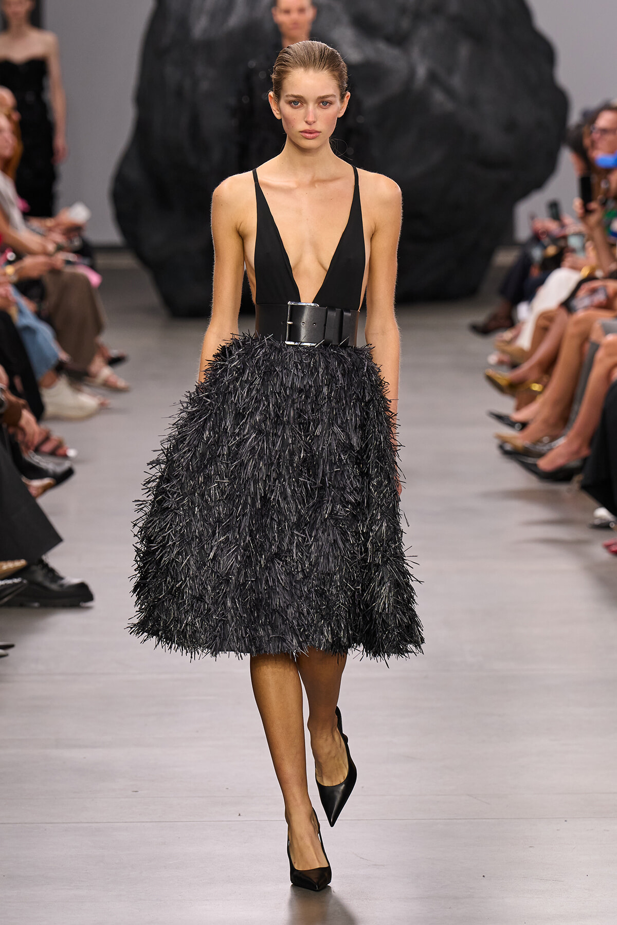 Model walks the runway in a black plunge-neck bodice and voluminous textured black skirt with a wide belt.