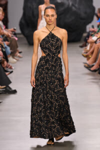 Model wearing a black halter-neck, cut-out floral lace evening gown on a runway, walking toward the camera.