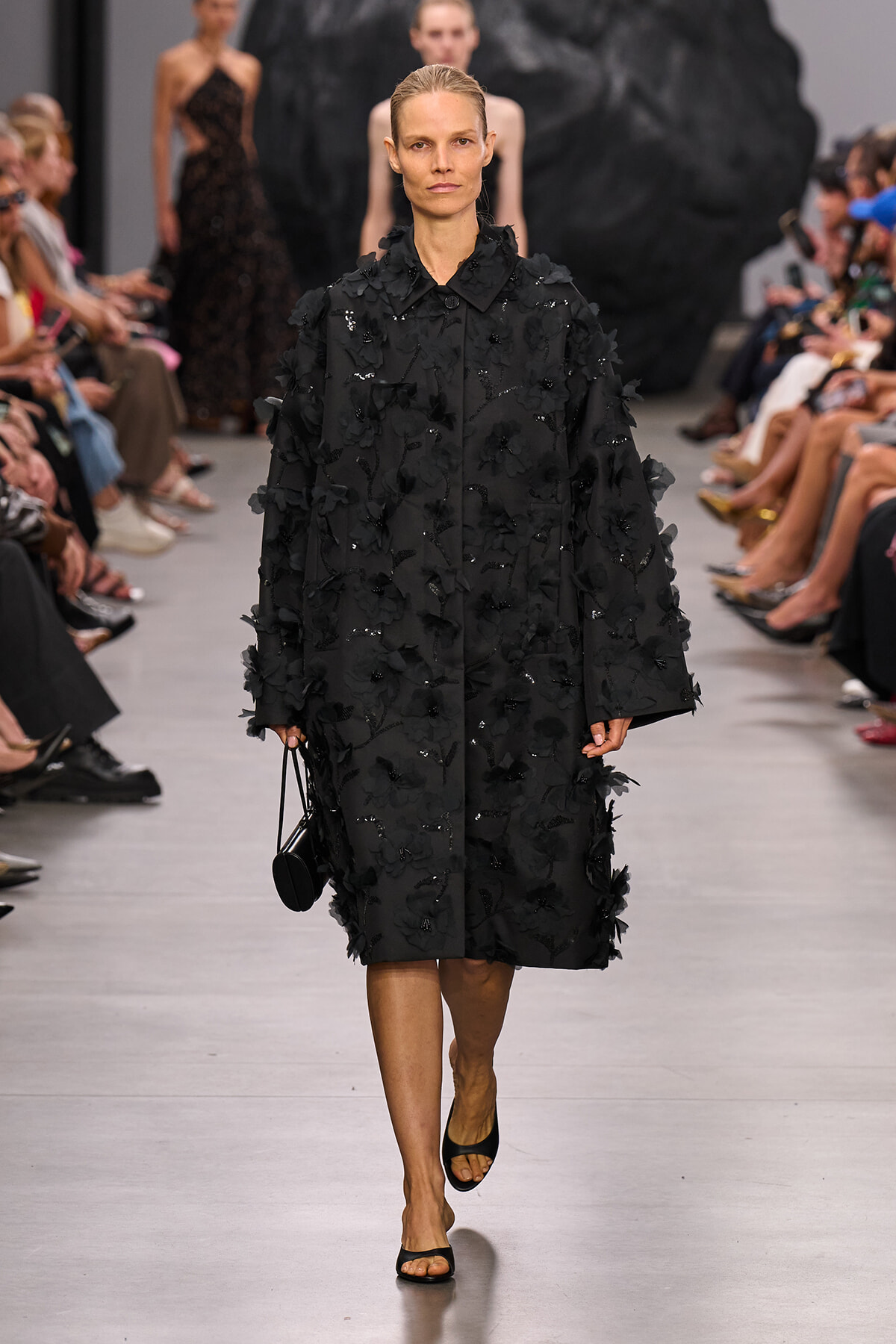 Model walks the runway in a long black coat covered in 3D floral appliqués, paired with black sandals and a small round bag.