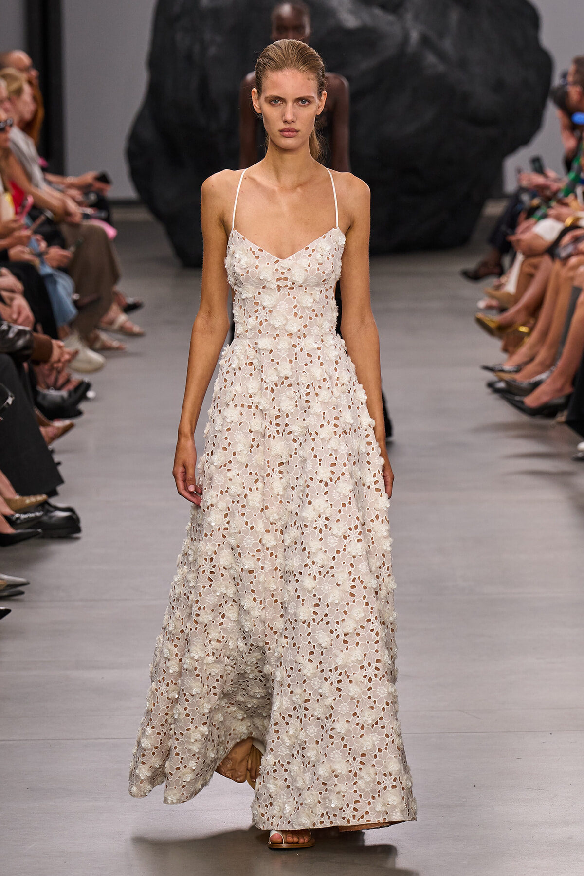 Runway model in a floor-length white floral lace dress with thin straps, walking the catwalk.