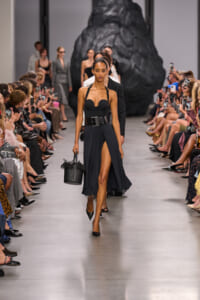 Model walks the runway in a black halter top and pleated high-slit skirt, carrying a woven black handbag; audience lines the catwalk behind.