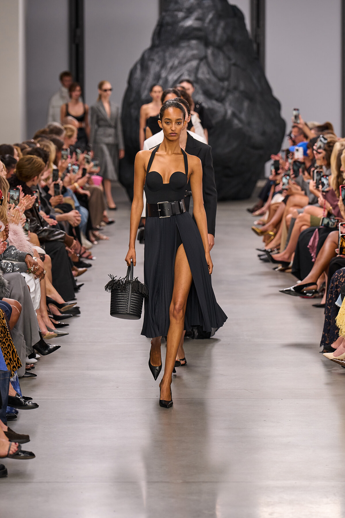 Model walks the runway in a black halter top and pleated high-slit skirt, carrying a woven black handbag; audience lines the catwalk behind.