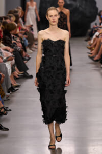 Model walks runway in a strapless black floral-embellished midi dress, audience on both sides watching.