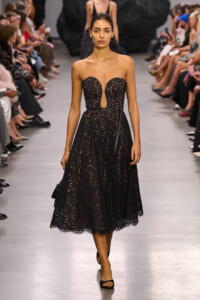 Model walks a runway in a black lace strapless midi dress with a peep-through bodice and belt, audience on both sides.