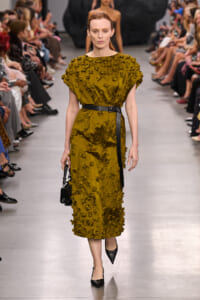 Model walks a fashion runway in a mustard yellow textured midi dress with floral appliqués, cinched with a black belt, carrying a small black handbag.