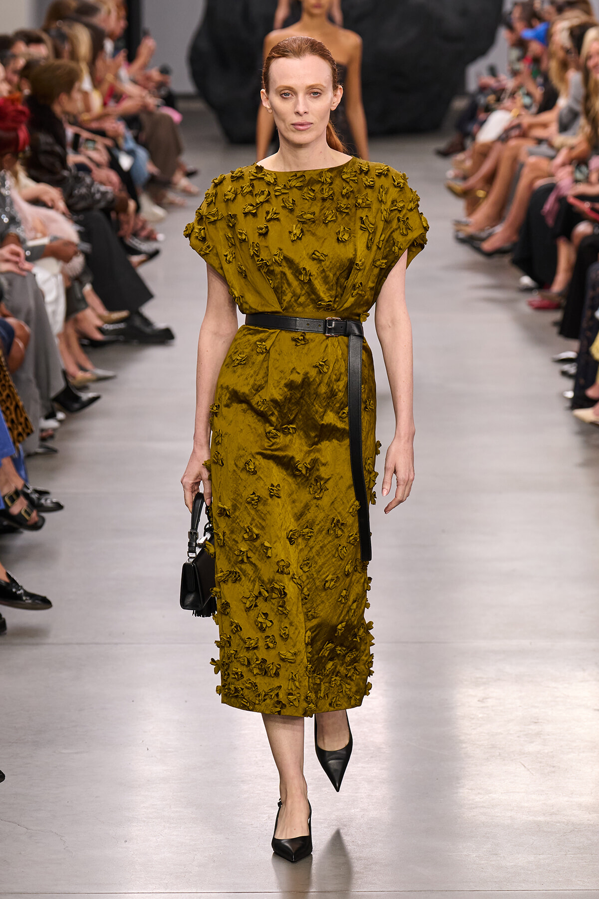 Model walks a fashion runway in a mustard yellow textured midi dress with floral appliqués, cinched with a black belt, carrying a small black handbag.