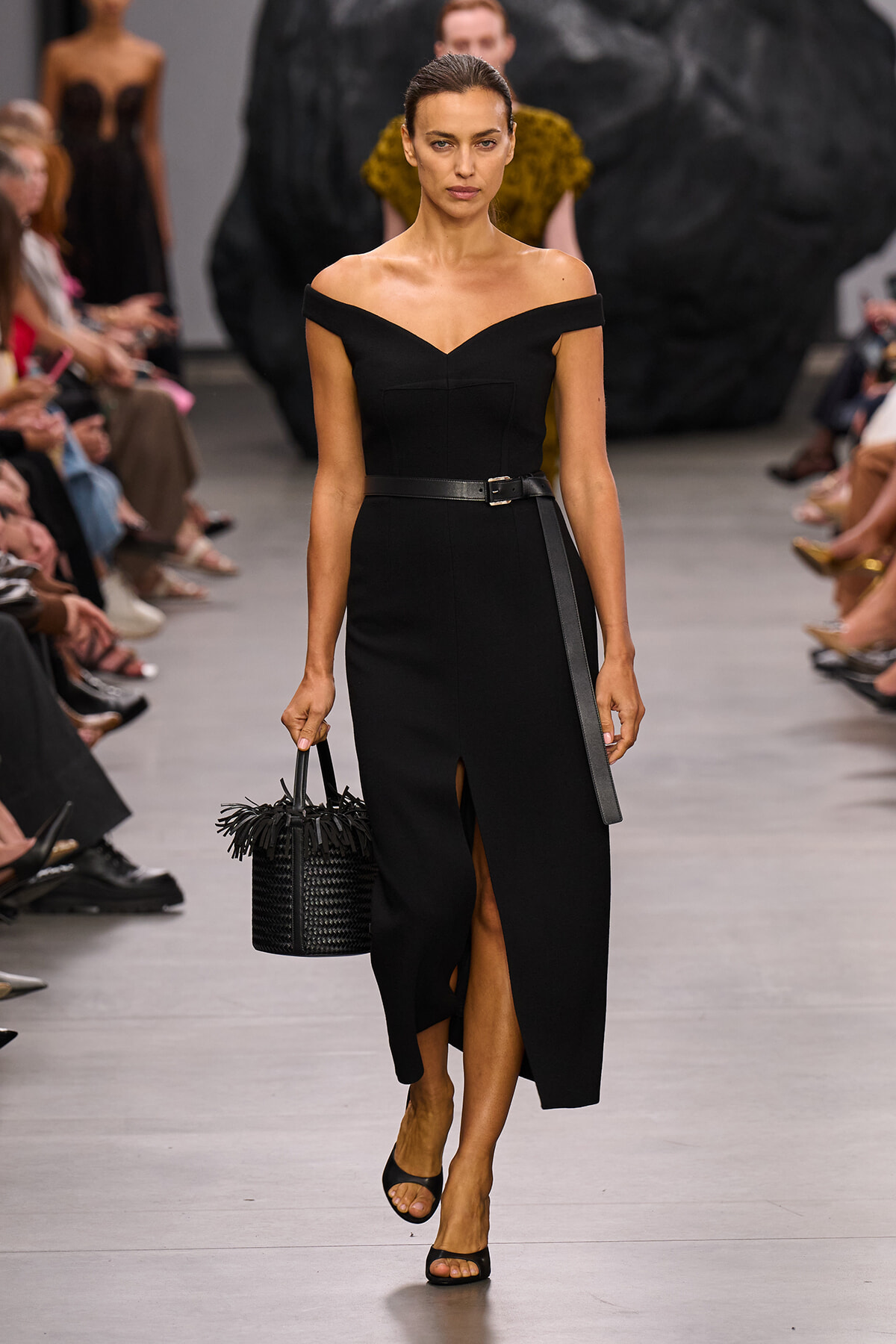 Model walks a black off-shoulder midi dress with belt, carrying a woven black handbag on a fashion runway.