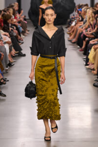 Model walks a fashion runway in a black button-down blouse tucked into a mustard-yellow textured midi skirt with floral appliqués, paired with a black belt and black fringe handbag.