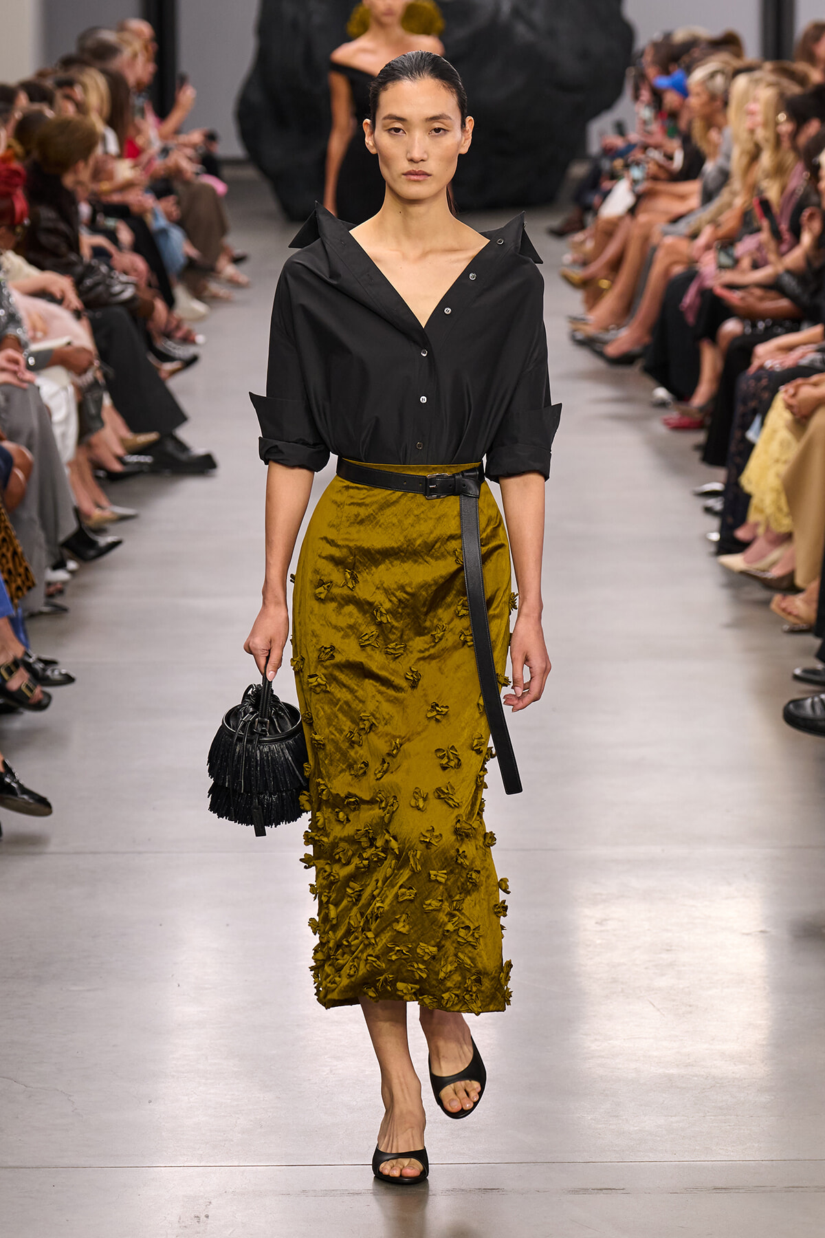 Model walks a fashion runway in a black button-down blouse tucked into a mustard-yellow textured midi skirt with floral appliqués, paired with a black belt and black fringe handbag.