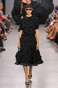 Model in a black knee-length dress covered with 3D floral appliqués walks the runway, wearing dark sunglasses.",