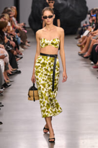 Model walks a fashion runway in a lime green and white strapless two-piece set with a black belt, sunglasses, and a straw handbag.