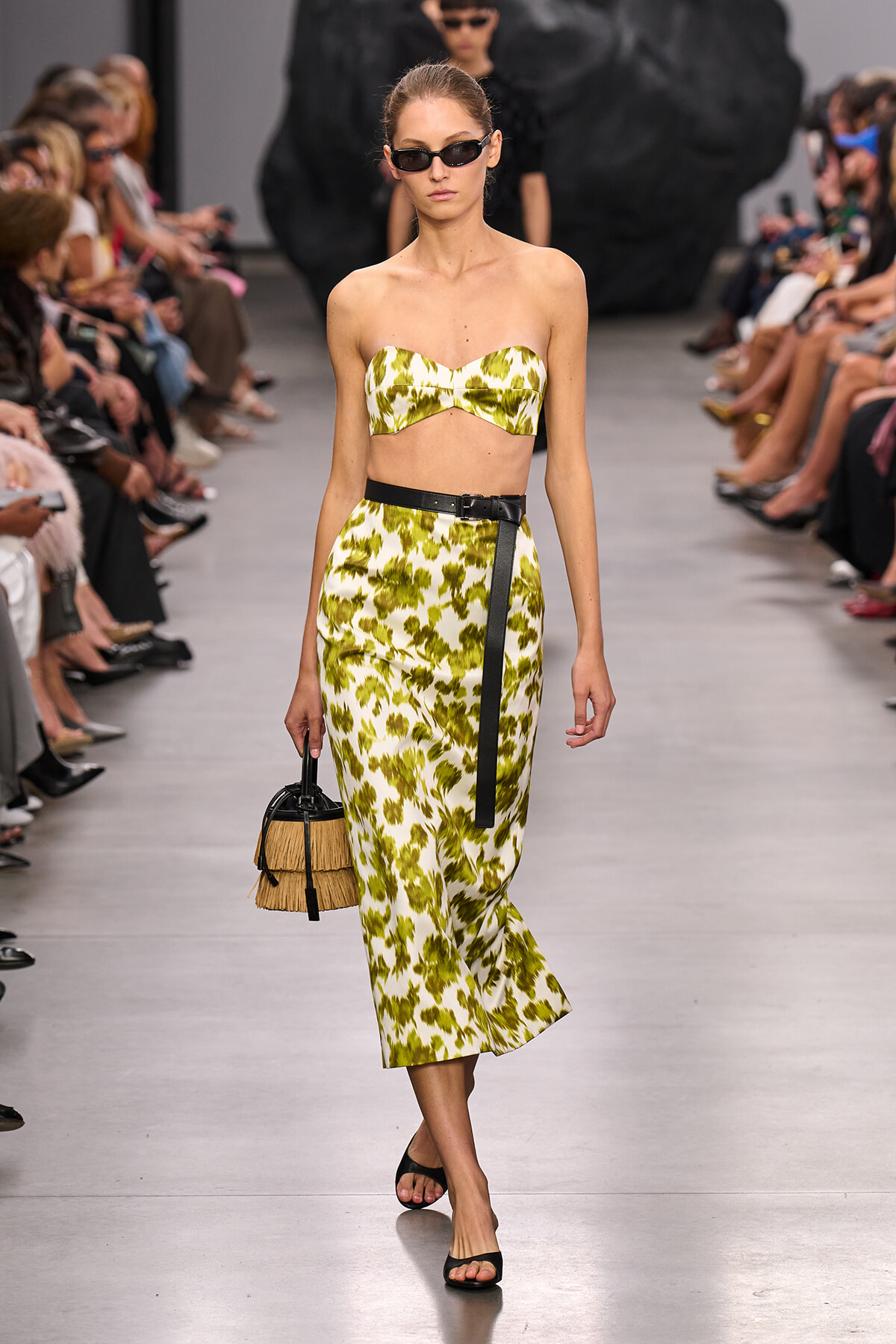 Model walks a fashion runway in a lime green and white strapless two-piece set with a black belt, sunglasses, and a straw handbag.