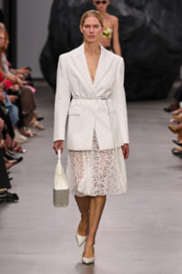 Model on a runway wearing a white belted blazer over a lace midi dress, carrying a white/gray handbag and wearing white heels.