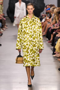 Model on a runway wearing a long yellow-green abstract print coat, black pointed heels, and a tan fringed handbag.