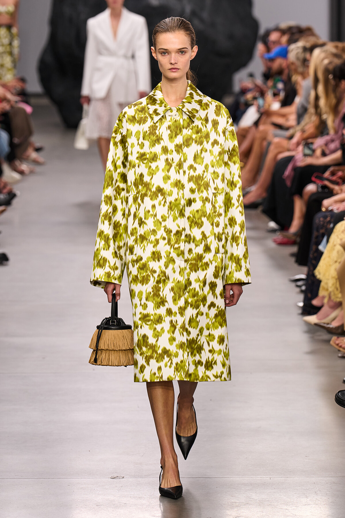 Model on a runway wearing a long yellow-green abstract print coat, black pointed heels, and a tan fringed handbag.