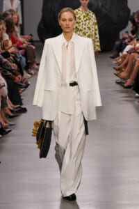 Model walks the runway in an all-white suit: oversized blazer, sheer blouse, and wide-leg trousers, carrying a black bag with gold tassels tassels on the left.