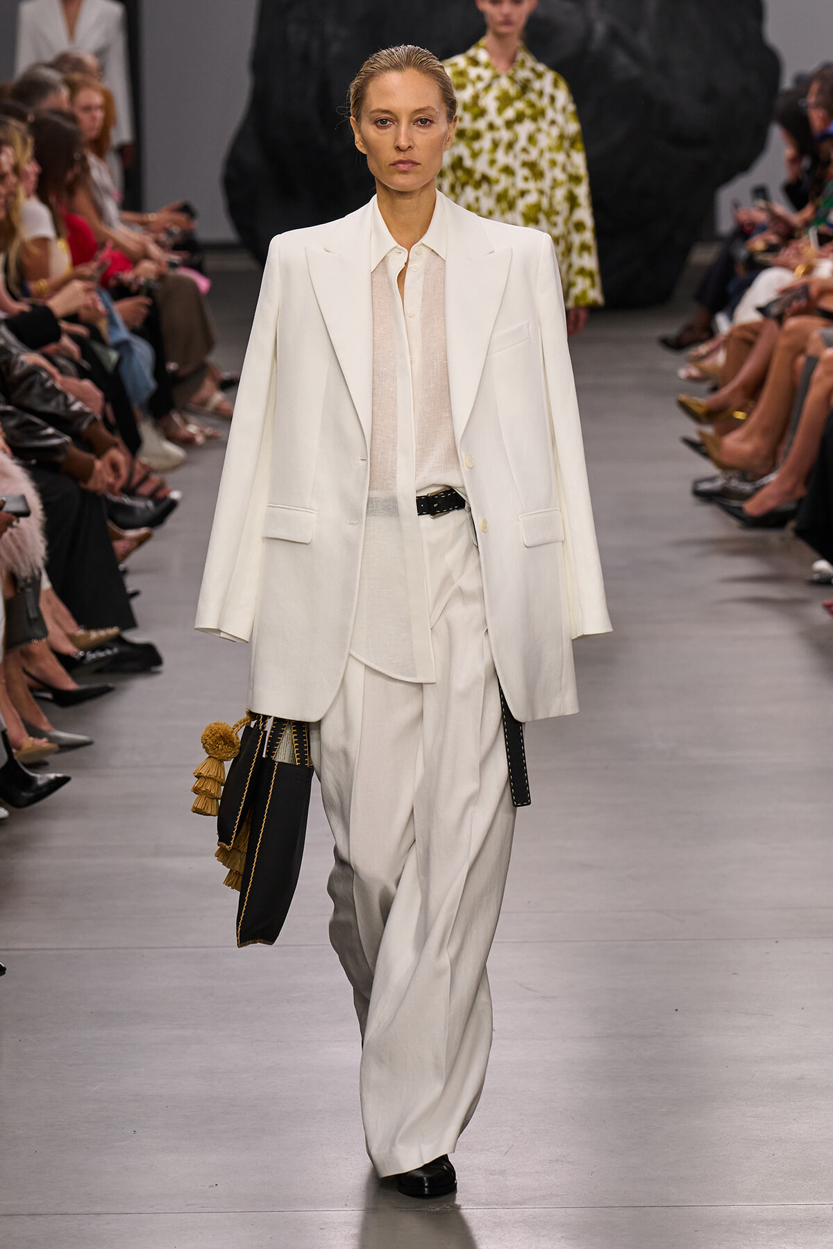 Model walks the runway in an all-white suit: oversized blazer, sheer blouse, and wide-leg trousers, carrying a black bag with gold tassels tassels on the left.