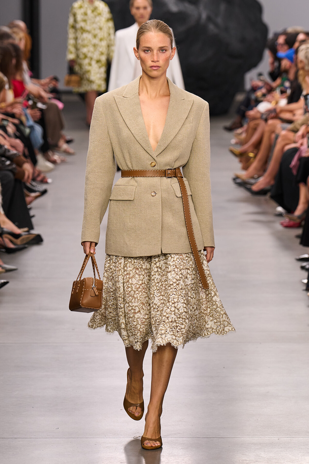 Model walks the runway in a beige blazer with a brown belt, paired with a lace midi skirt and tan sandals, carrying a small brown handbag