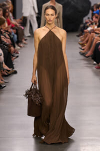 Model walks a runway in a brown halter-neck, pleated gown, carrying a brown woven tote with fringe at the side.
