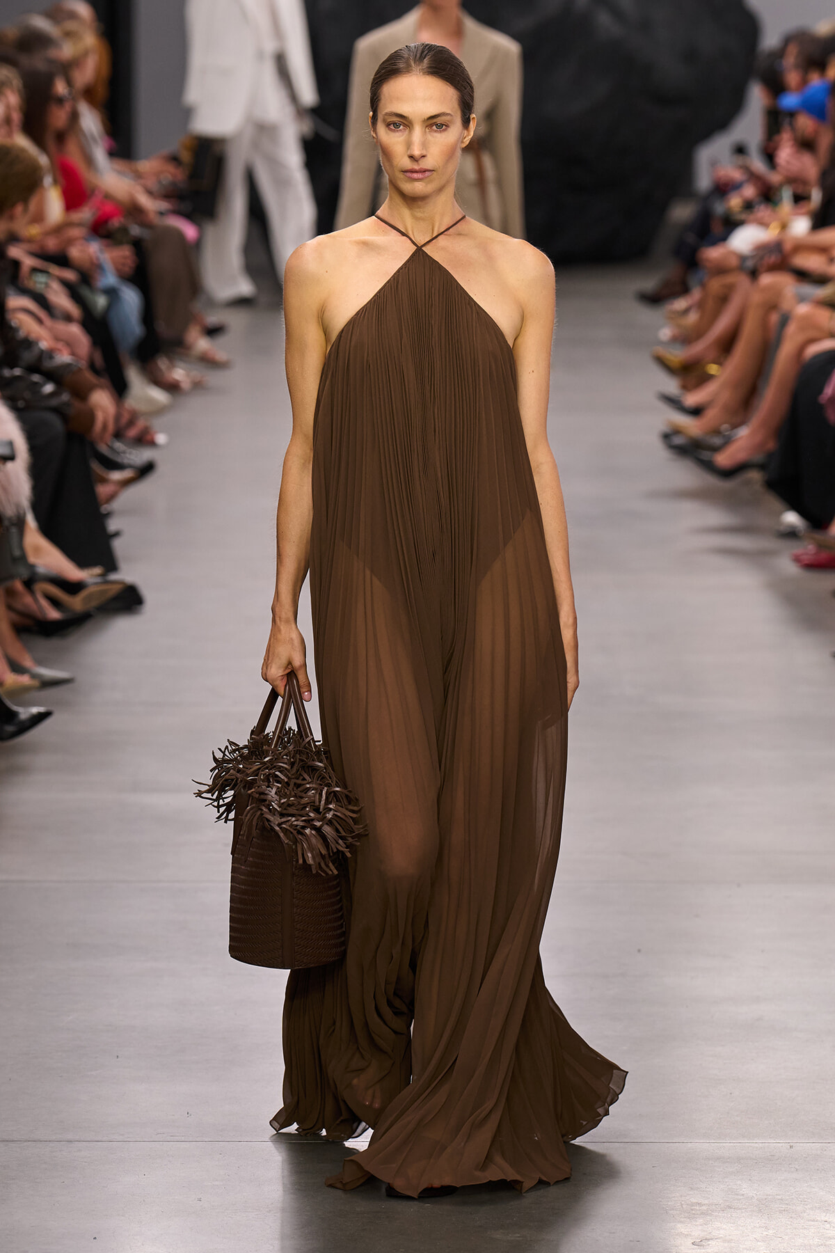 Model walks a runway in a brown halter-neck, pleated gown, carrying a brown woven tote with fringe at the side.