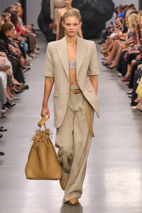 Model walking the runway in a beige suit with a cropped top, holding a large woven tan handbag.