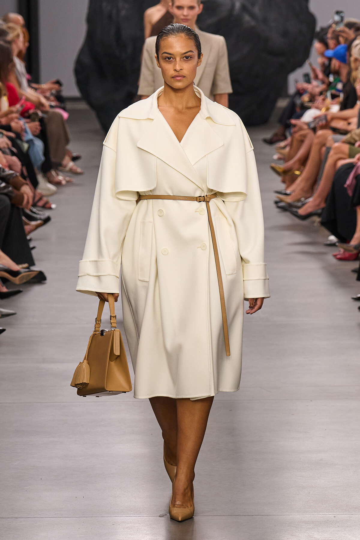 Model on a runway wearing a white belted trench coat with wide lapels, carrying a tan structured handbag and nude pumps.