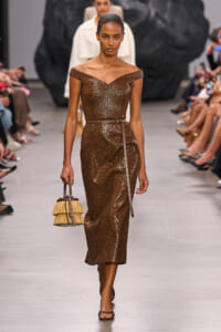 Model walking the runway in a brown sequin off-shoulder midi dress with a slim belt, carrying a fringed straw handbag.