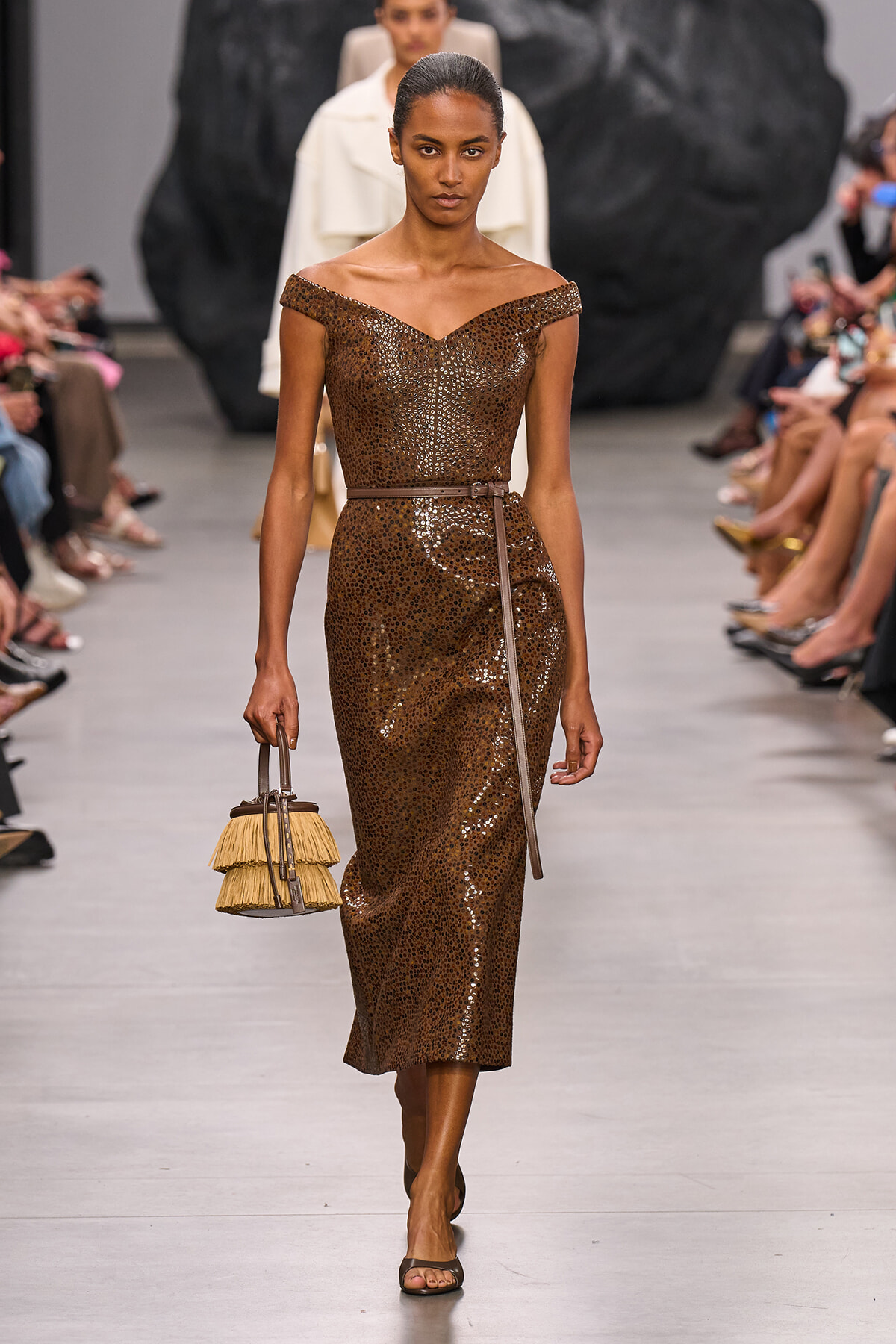 Model walking the runway in a brown sequin off-shoulder midi dress with a slim belt, carrying a fringed straw handbag.