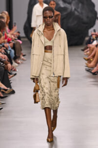 Model walks runway in beige windbreaker, matching cropped top and embroidered midi skirt, tan heels, and brown handbag with sunglasses.
