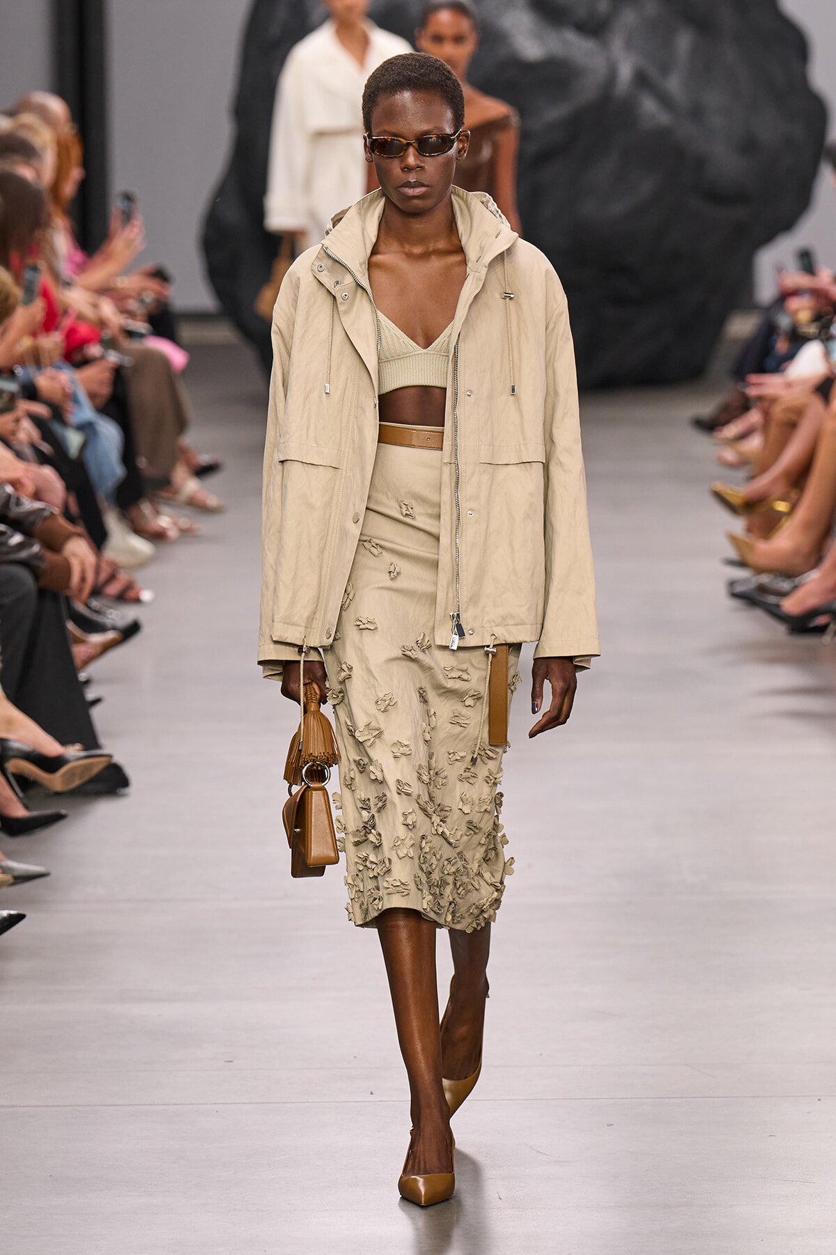 Model walks runway in beige windbreaker, matching cropped top and embroidered midi skirt, tan heels, and brown handbag with sunglasses.