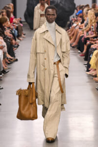 Male model on a runway wearing a beige trench coat over a light gray turtleneck, tan belt, and wide-leg pants, carrying a brown woven handbag.