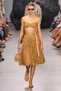 Model on a runway in a tan lace bandeau top and knee-length lace skirt with a tan belt and nude sandals, wearing sunglasses.