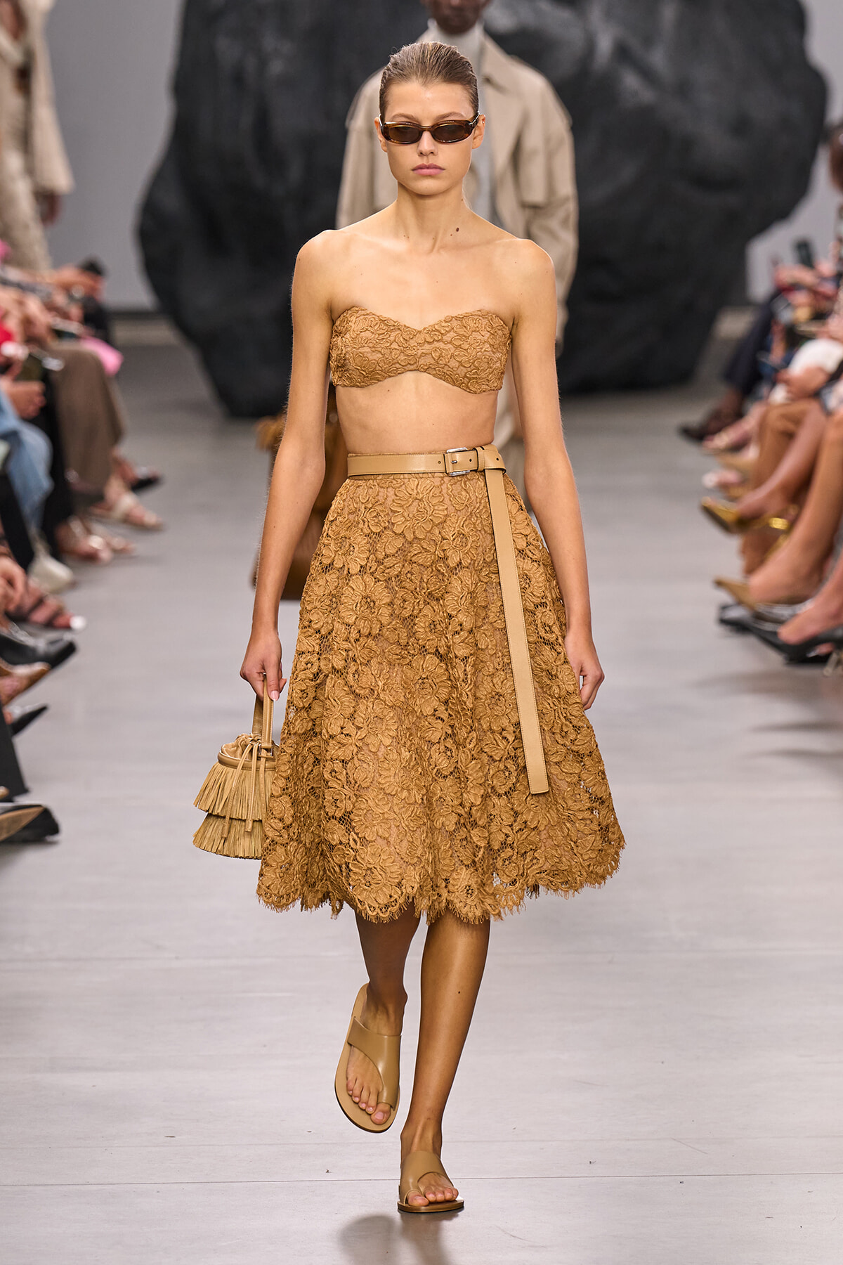 Model on a runway in a tan lace bandeau top and knee-length lace skirt with a tan belt and nude sandals, wearing sunglasses.